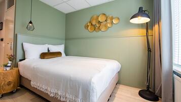 A neatly made bed with a white duvet and a brown pillow, a wooden nightstand with a plant, and a wall-mounted light fixture.
