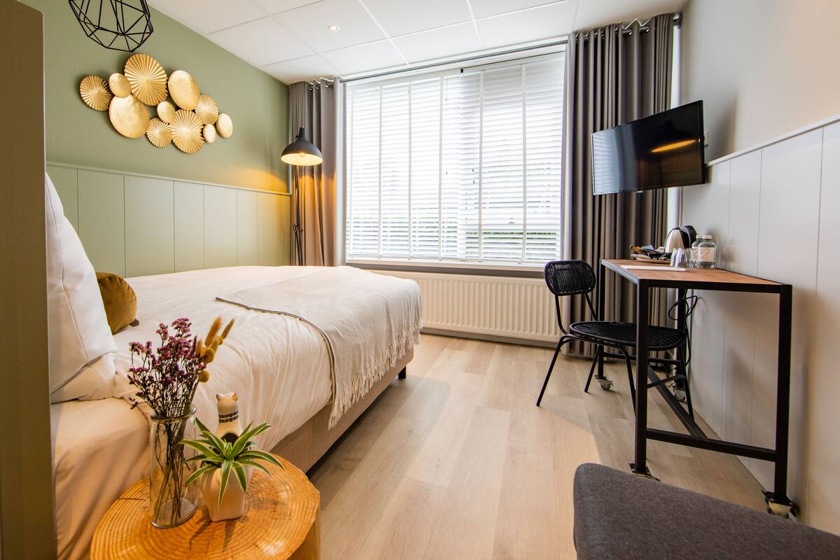 A hotel room with a bed, a desk with a chair, a TV, and a window with curtains.