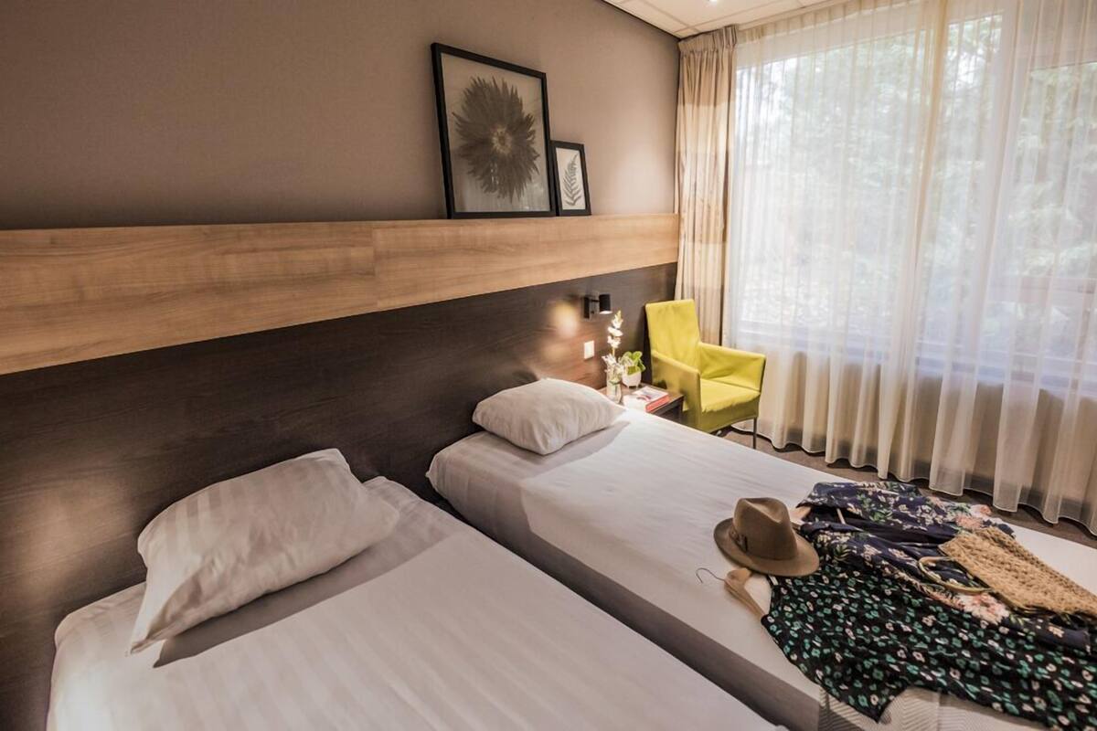 A hotel room with two beds, a wooden headboard, a yellow chair, and a large window with sheer curtains.