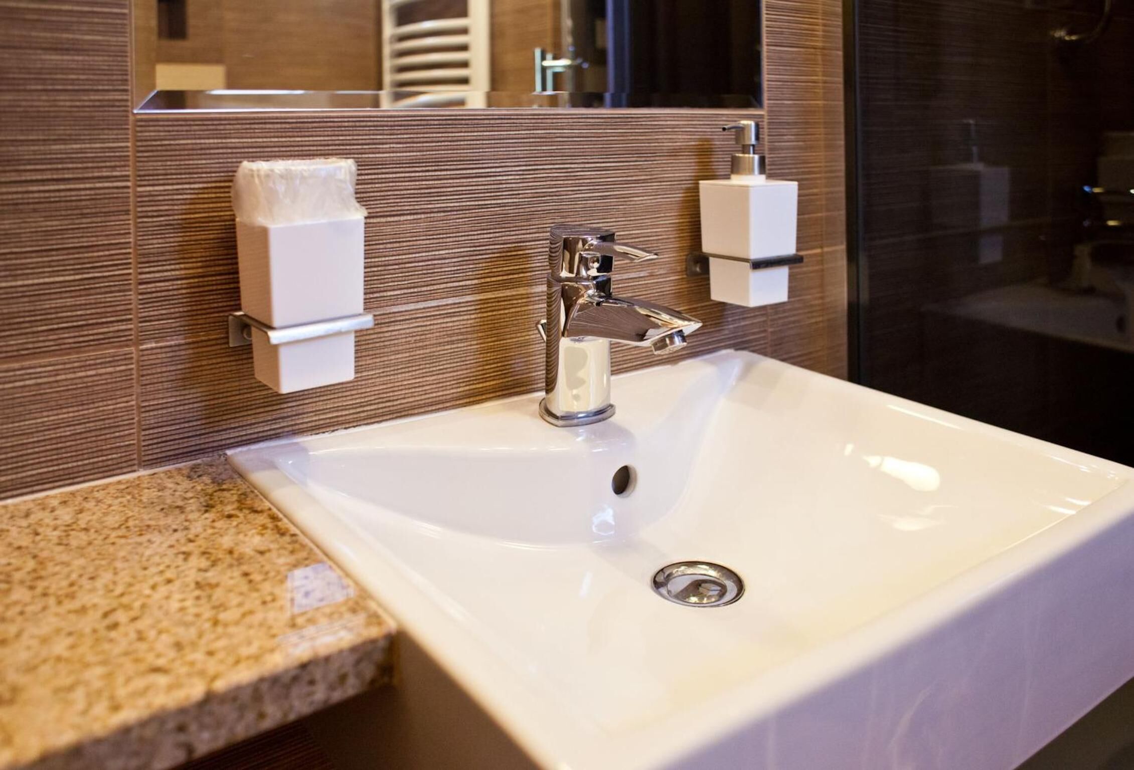 A bathroom with a white sink, chrome faucet, and a mirror above it. There are two soap dispensers on the wall.
