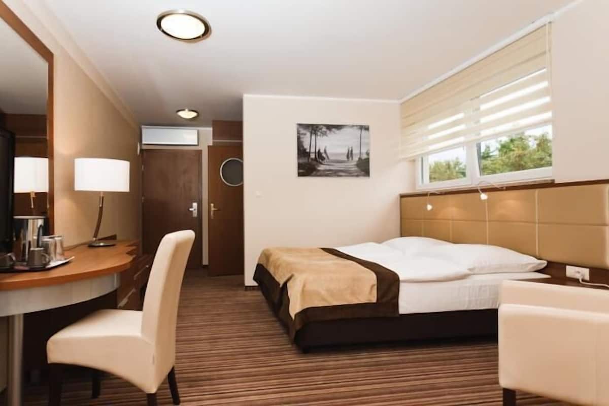 A hotel room with a bed, a desk with a chair, a TV, and a window with blinds.
