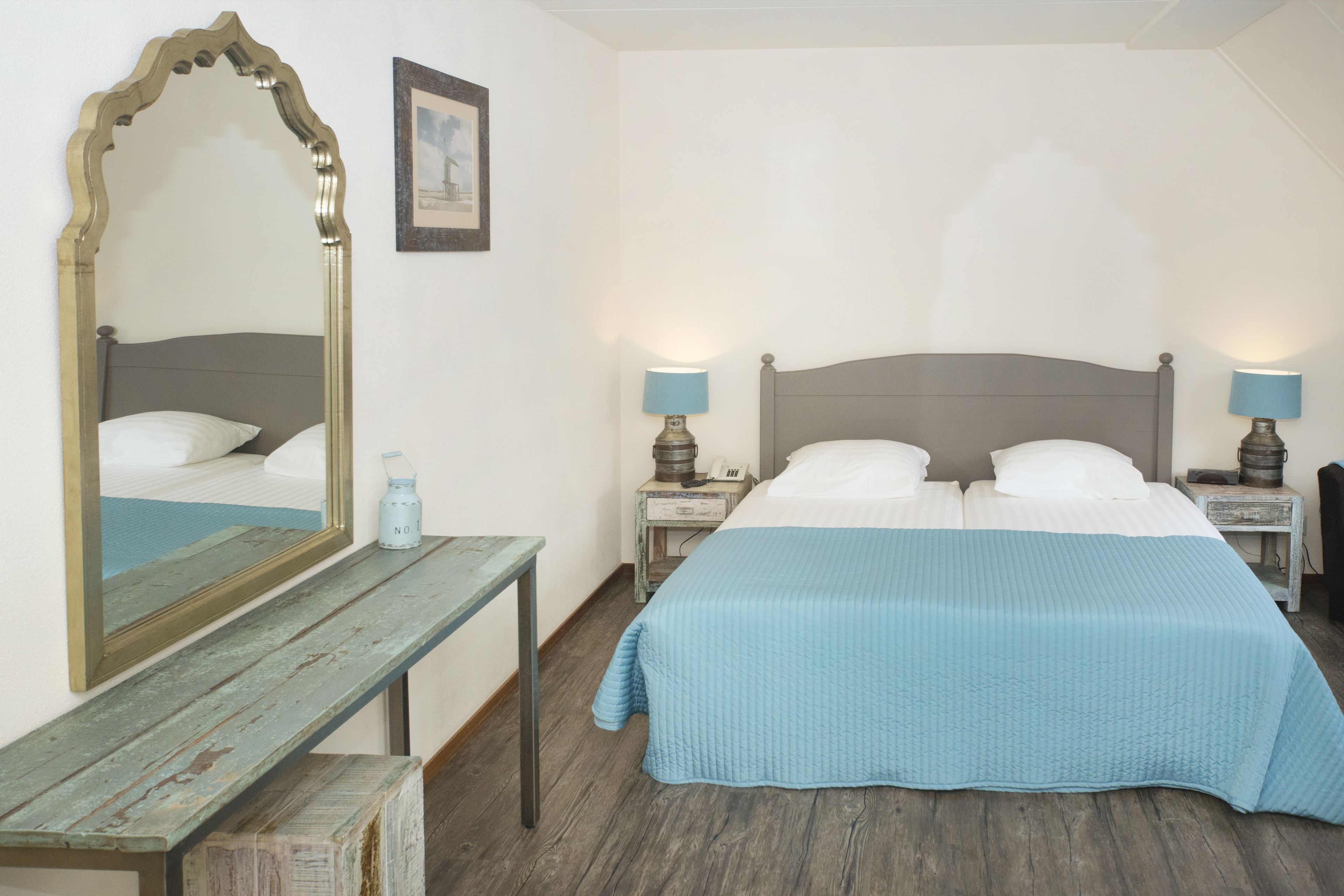 A bedroom with a large mirror, a bed with two pillows, a bedside table with a lamp, and a wooden bench.