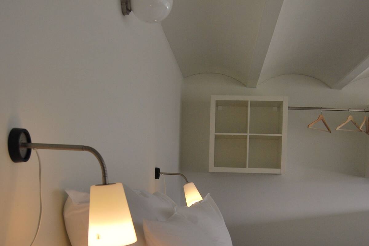Une chambre avec deux lits, chacun muni d’une lampe de chevet. Il y a une fenêtre donnant sur l’extérieur, ainsi qu’un porte-manteau avec des cintres.