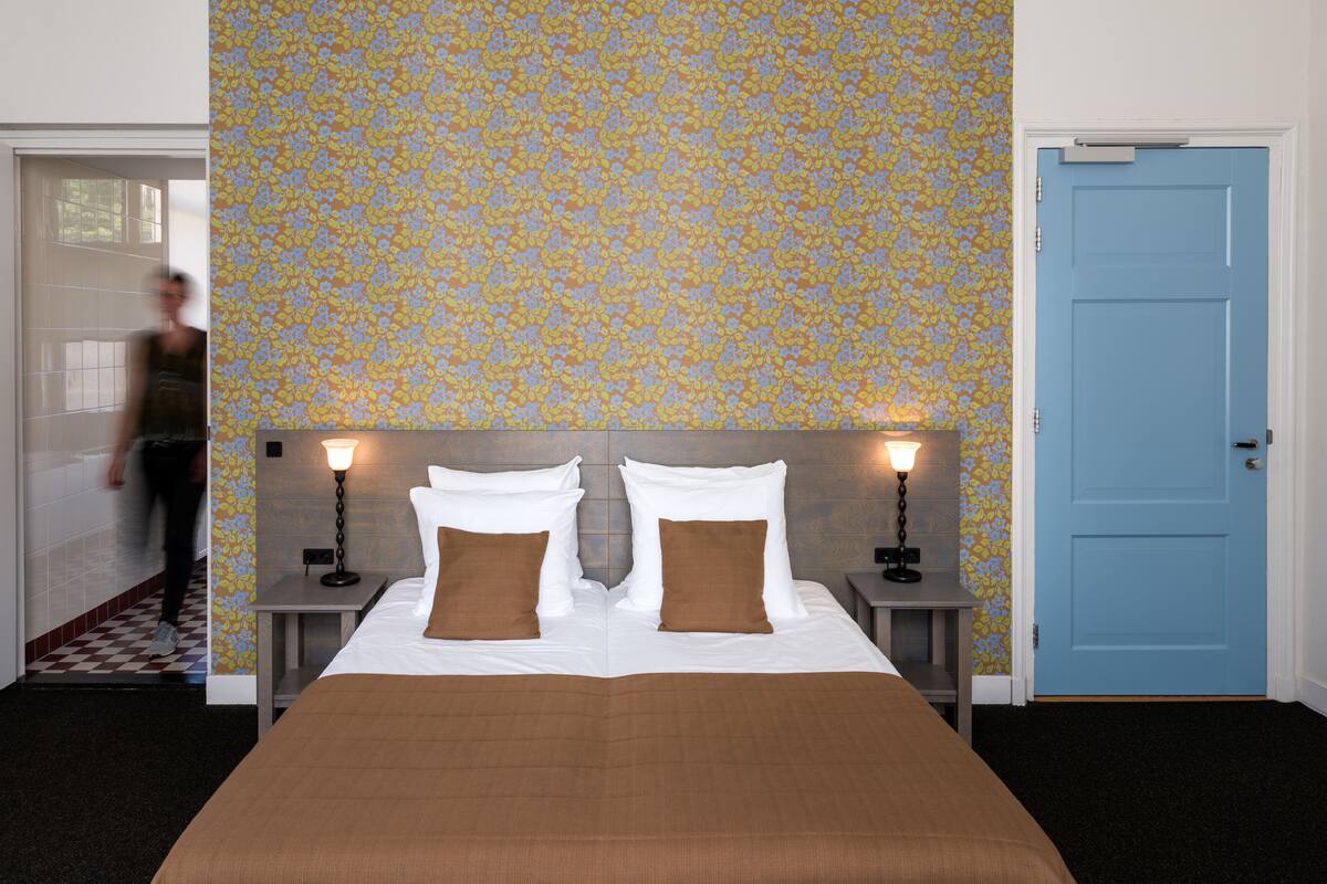 Une chambre d’hôtel avec un grand lit, deux tables de chevet avec des lampes, une porte bleue et un papier peint à motifs.