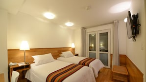 Ein Hotelzimmer mit zwei Betten, einem Schreibtisch mit Lampe, einem an der Wand befestigten Fernseher und einer Glasschiebetür, die auf einen Balkon führt.
