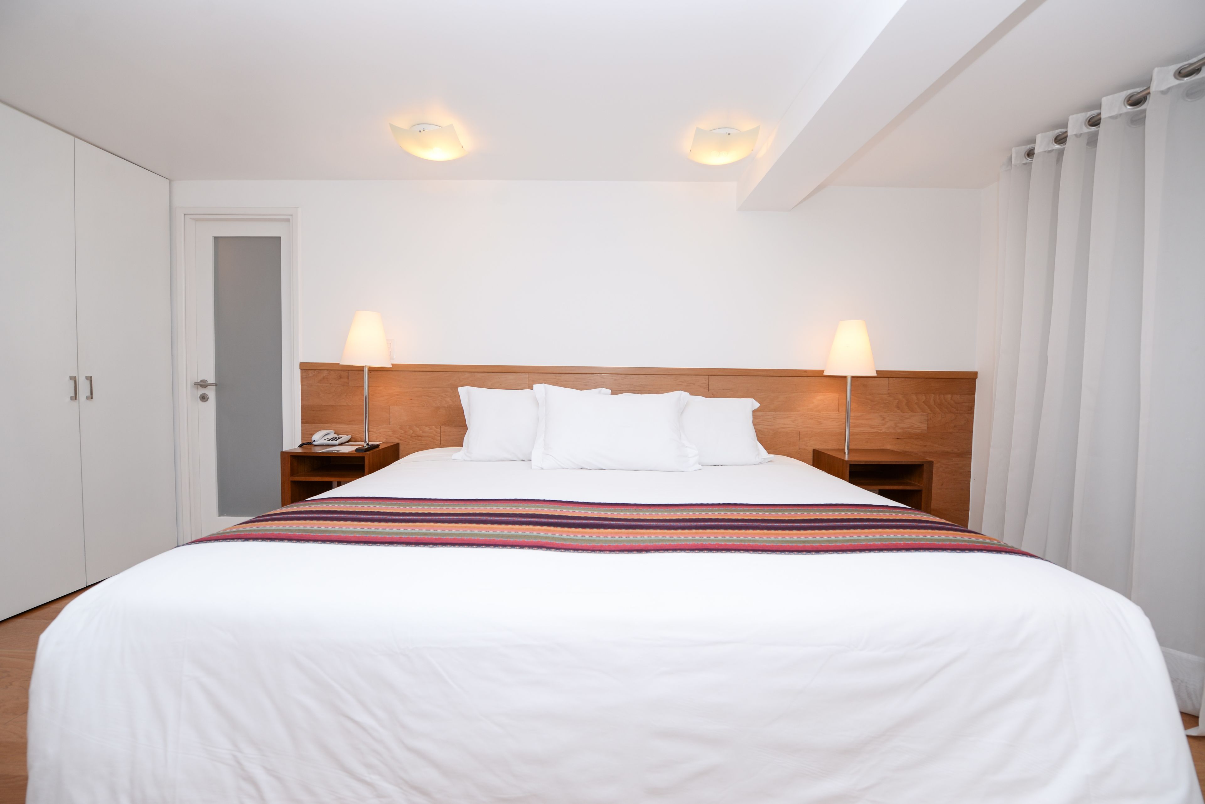 Une chambre d’hôtel avec un grand lit, du linge de lit blanc, deux lampes de chevet, une tête de lit en bois et une porte.