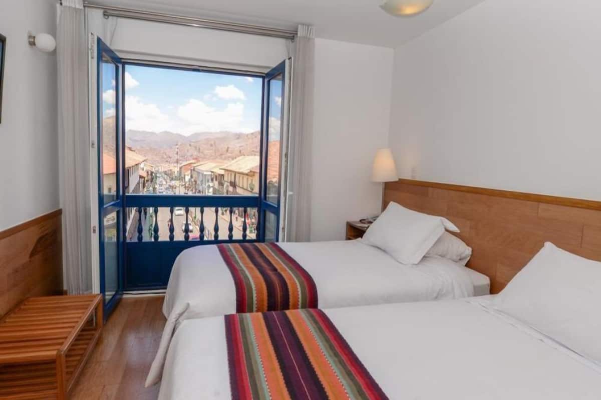 Habitación de hotel con dos camas, un balcón y vistas a la ciudad, además de un banco de madera.