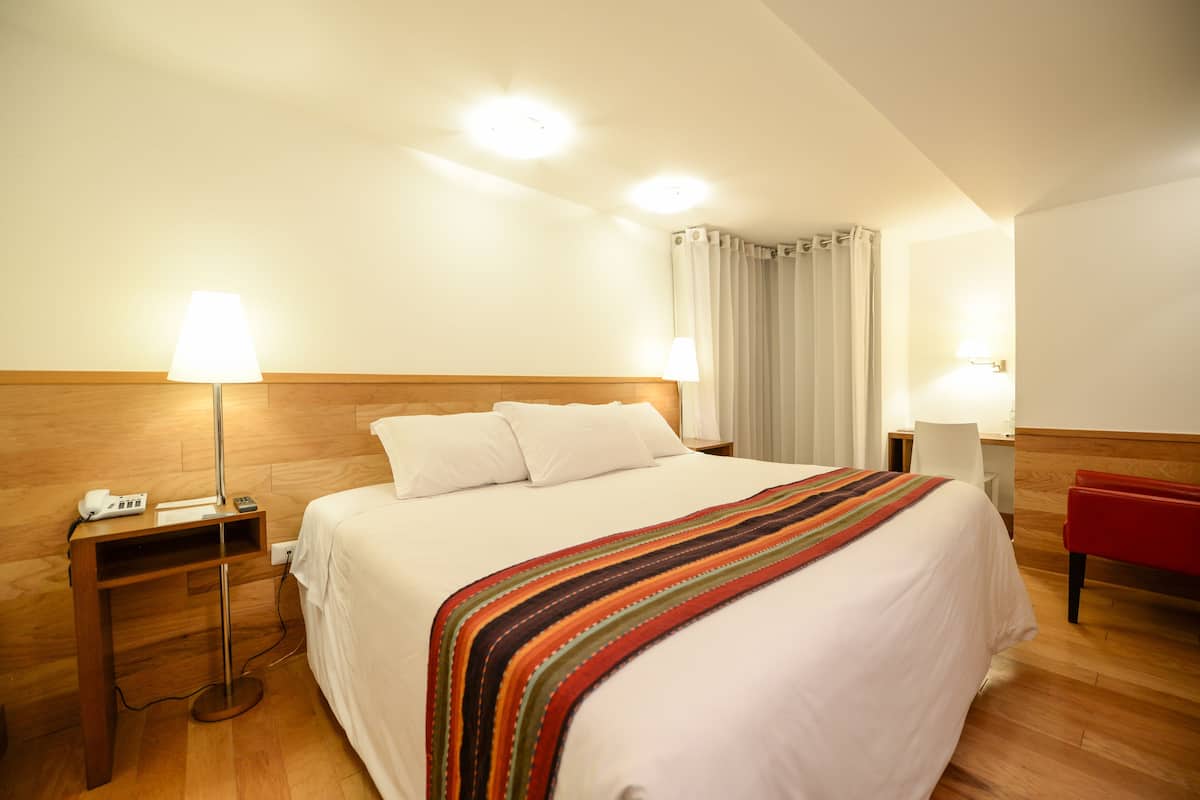 Habitación de hotel con una cama grande, dos mesitas de noche con lámparas, un banco rojo y un cabecero de madera.
