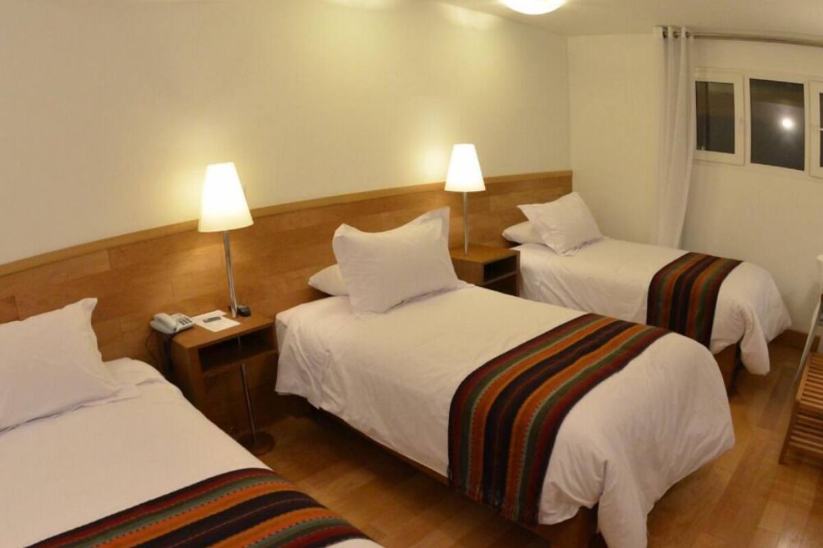 Habitación de hotel con tres camas individuales, cada una con una manta a rayas, una mesita de noche con lámpara y teléfono. El piso es de madera y las paredes son blancas.