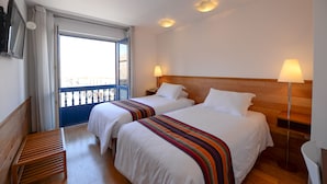 Ein Hotelzimmer mit zwei Betten, einer Holzbank, einem Balkon und einem an der Wand montierten Fernseher.
