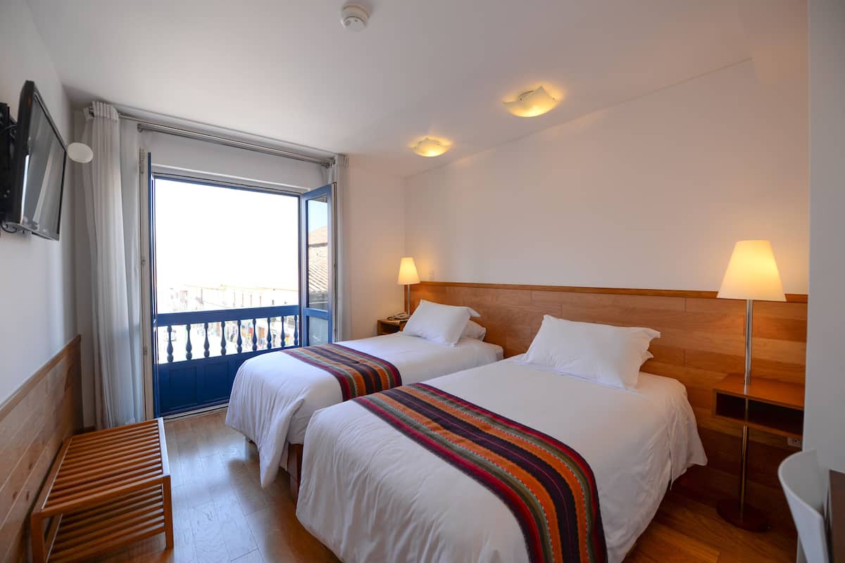 Habitación de hotel con dos camas, un banco de madera, un balcón y un televisor montado en la pared.