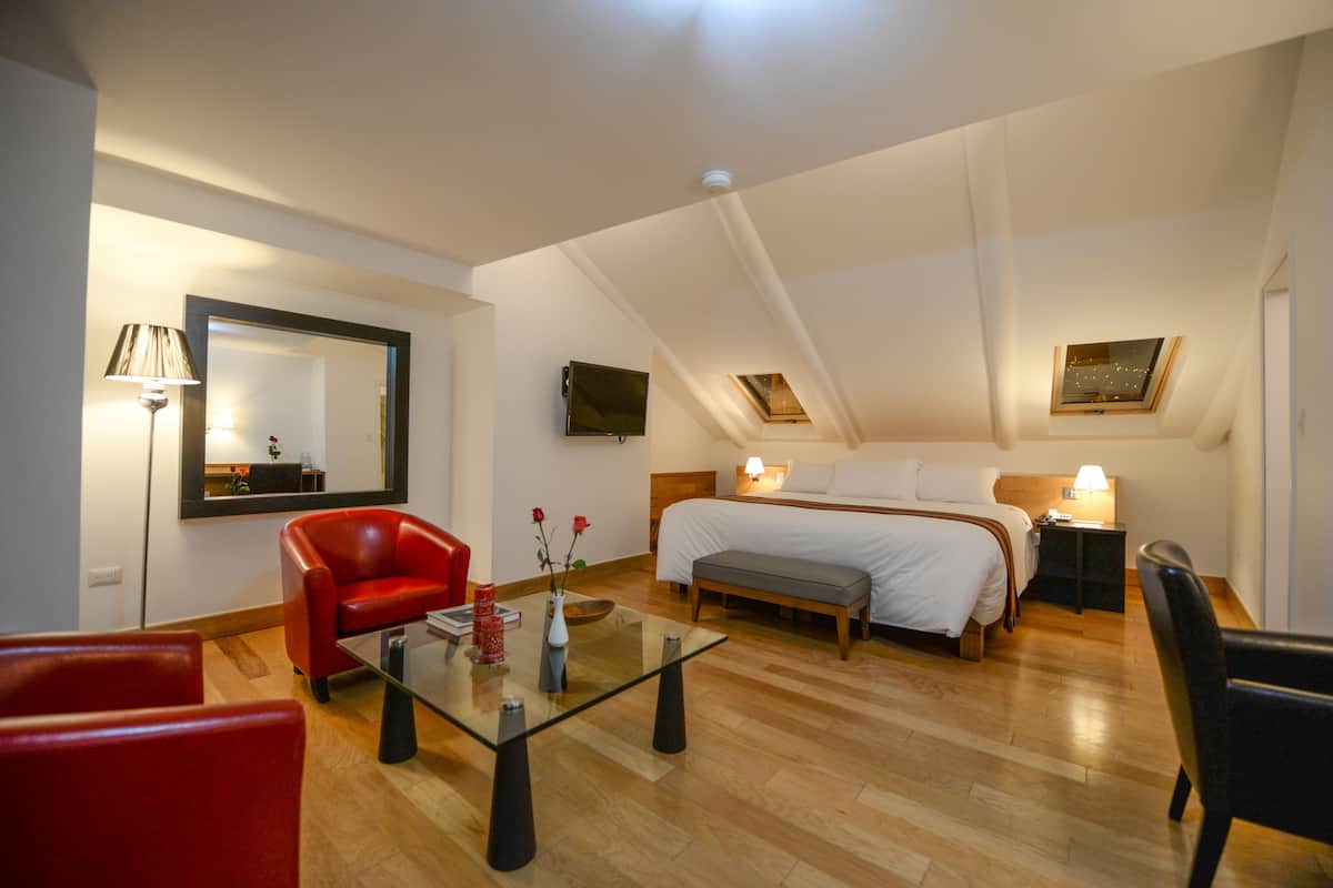 Habitación de hotel moderna con una cama grande, una zona de estar con una mesa de vidrio, un sillón rojo y un banco.