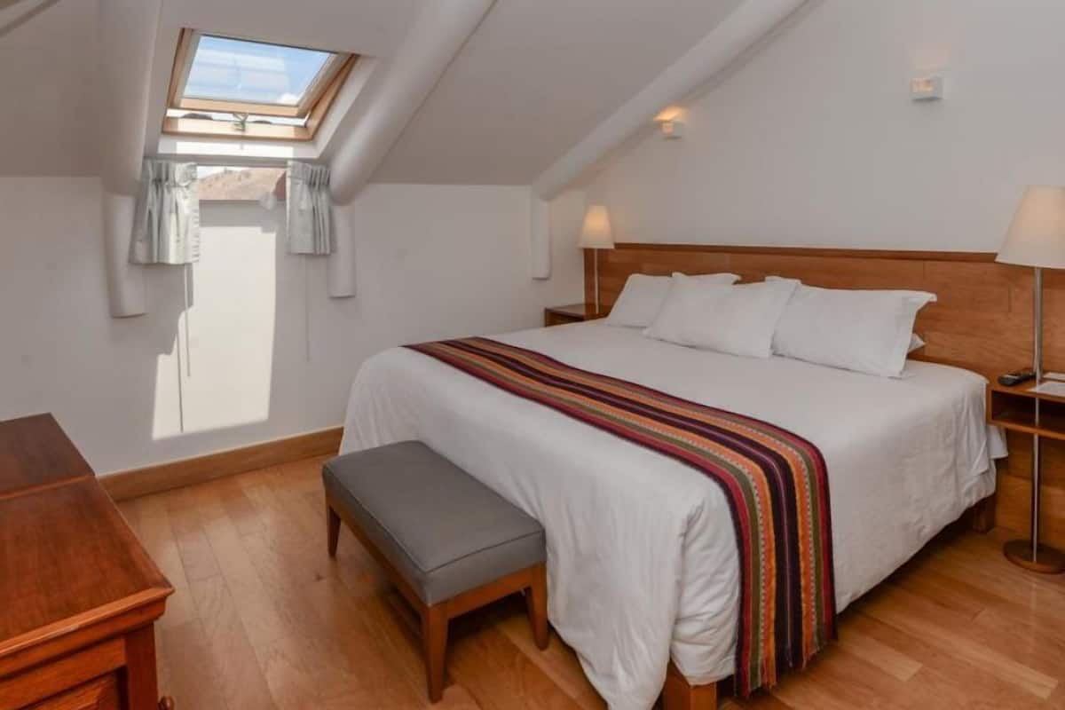 Un dormitorio con techo inclinado, tragaluz, cama con ropa de cama blanca, una banca y una mesita de noche.