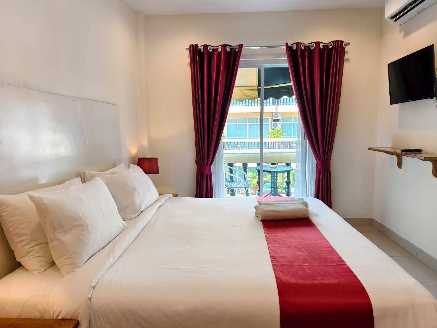 A hotel room with a bed, a red and white striped curtain, a TV mounted on the wall, and a balcony visible through the window.