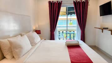 A hotel room with a bed, a red and white striped curtain, a TV mounted on the wall, and a balcony visible through the window.