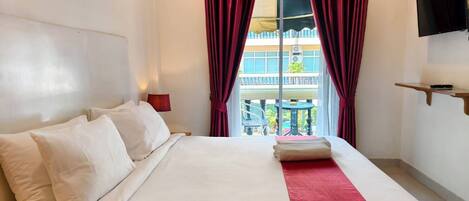 A hotel room with a bed, a red and white striped curtain, a TV mounted on the wall, and a balcony visible through the window.