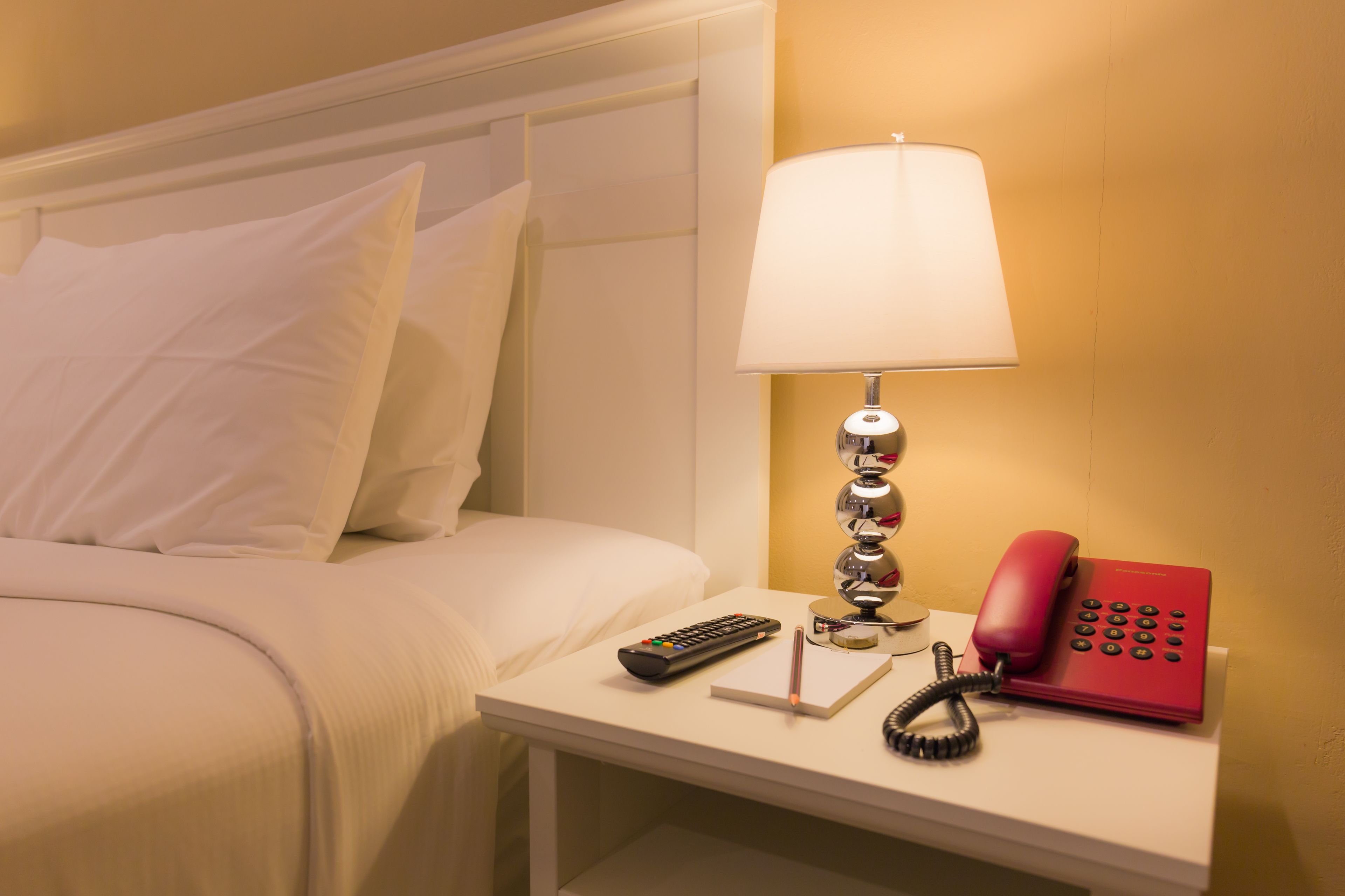 A neatly made bed with white linens, a bedside table with a lamp, a red telephone, and a remote control.