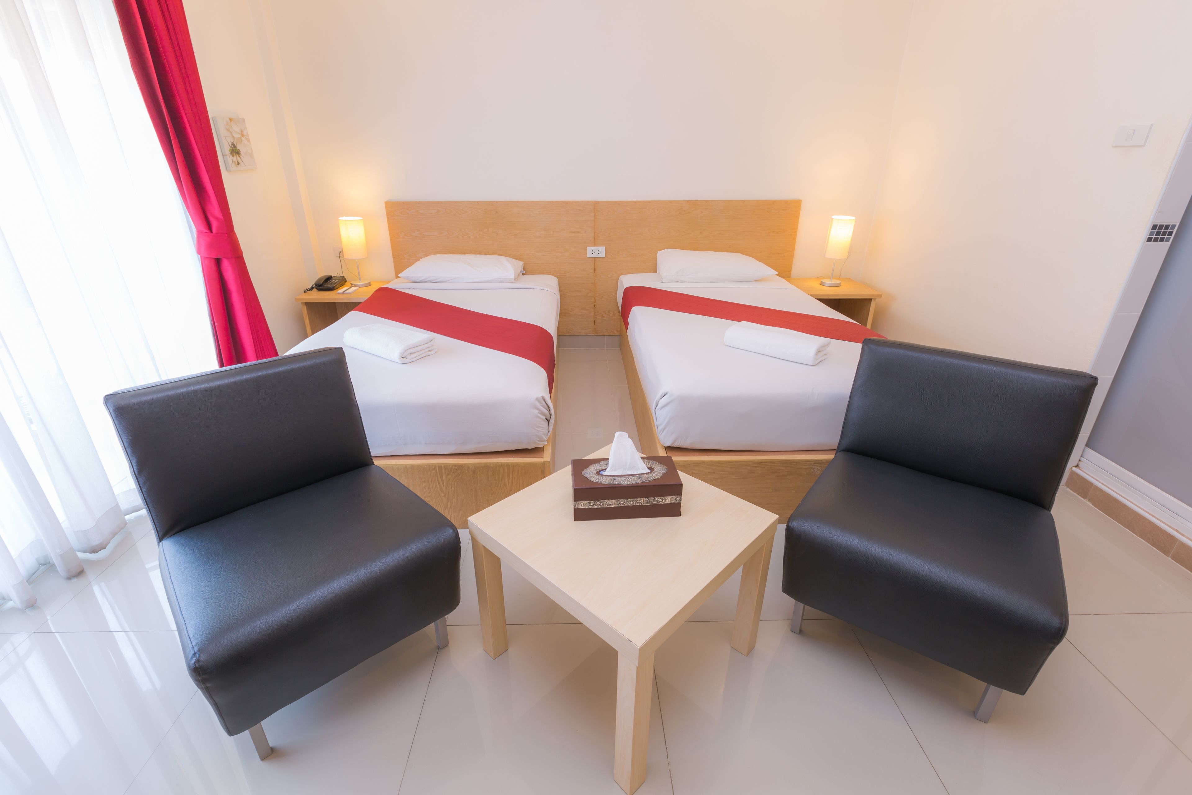 A hotel room with two beds, a small table, two black armchairs, and a red curtain.