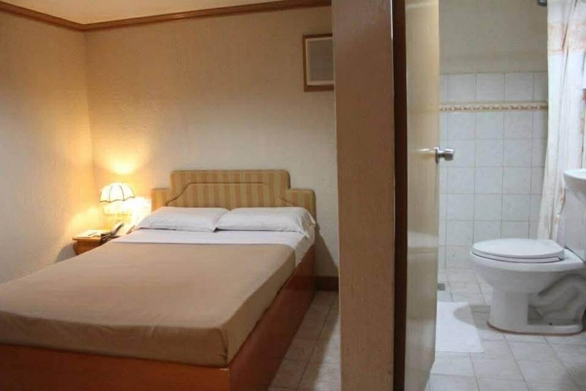 A hotel room with a bed, bedside lamp, and a view into the bathroom with a toilet and shower.
