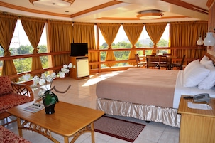 A hotel room with a bed, a desk, a TV, a dining area with chairs, and a balcony with curtains.