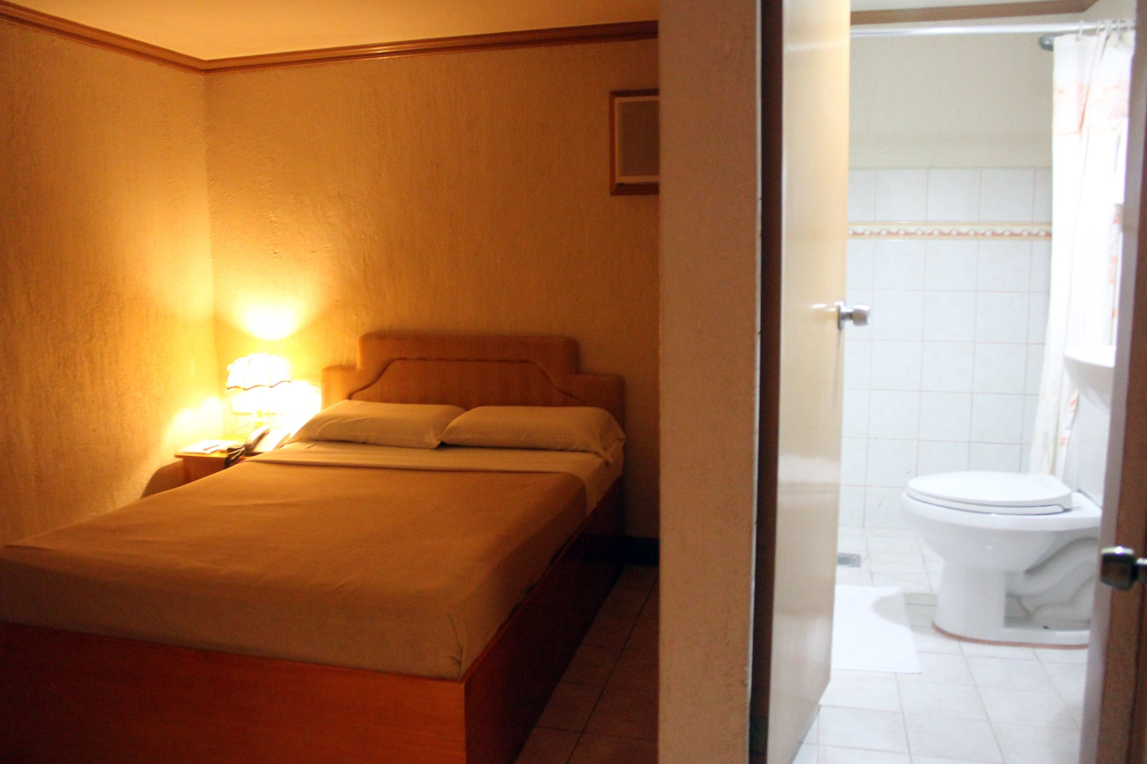 A hotel room with a bed, a nightstand with a lamp, and a bathroom with a toilet and shower.