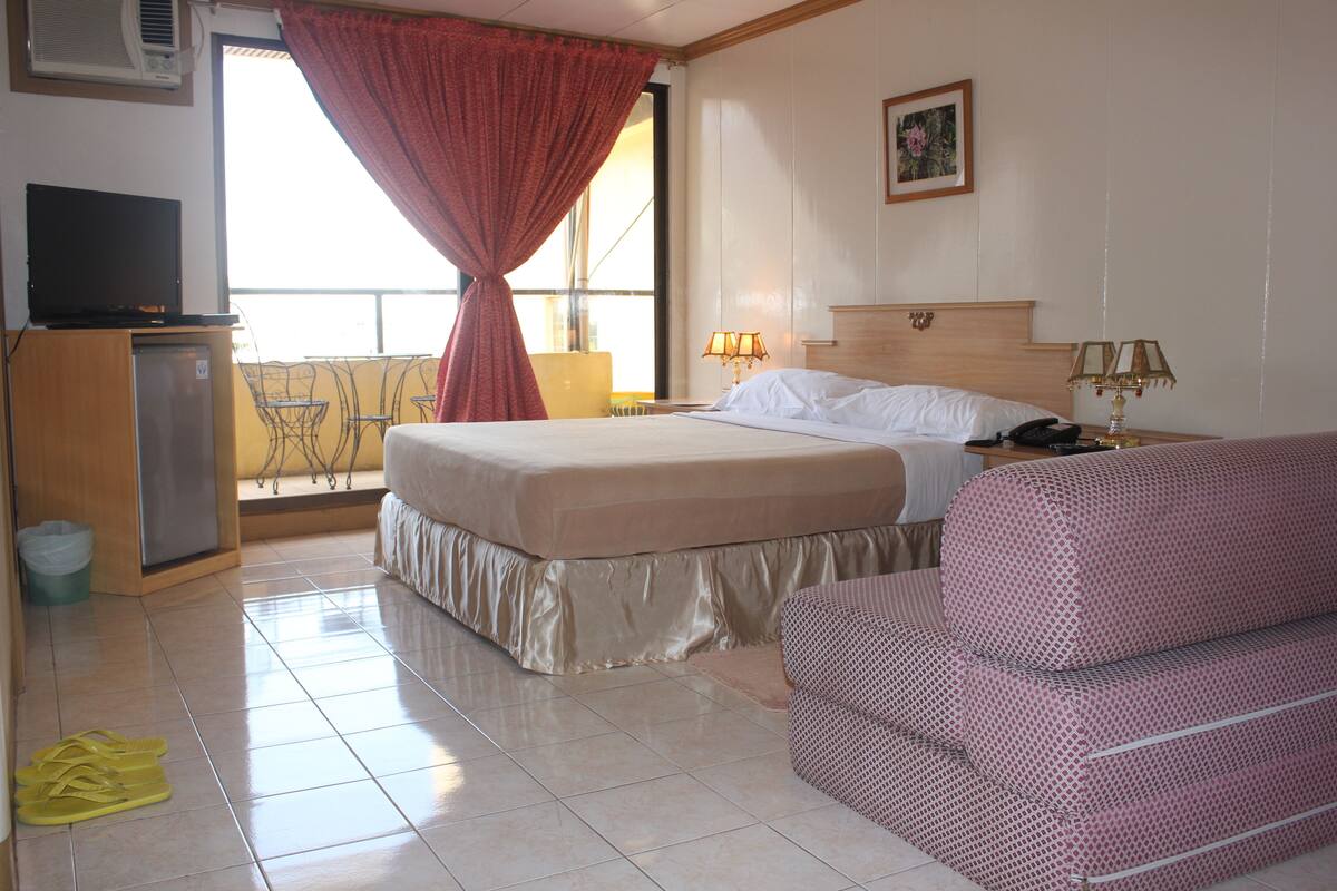 A hotel room with a bed, a sofa, a TV, a desk, and a balcony with a chair.