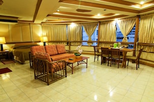 A hotel room with a sofa, dining table, chairs, a bed, and large windows with curtains.