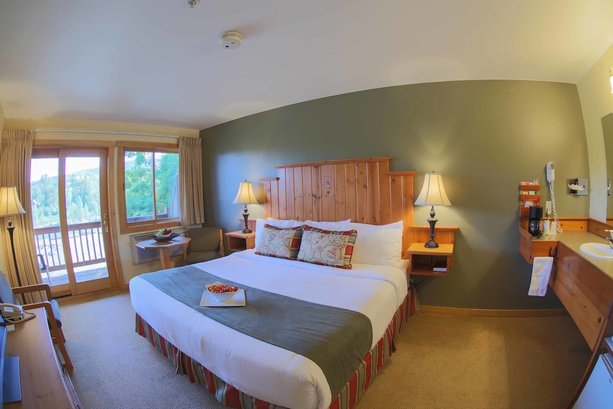 A hotel room with a large bed, two bedside tables with lamps, a wooden headboard, a small table with a bowl of fruit, and a view of the outdoors through a sliding door.