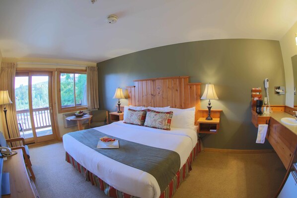 A hotel room with a large bed, two bedside tables with lamps, a wooden headboard, a small table with a bowl of fruit, and a view of the outdoors through a sliding door.