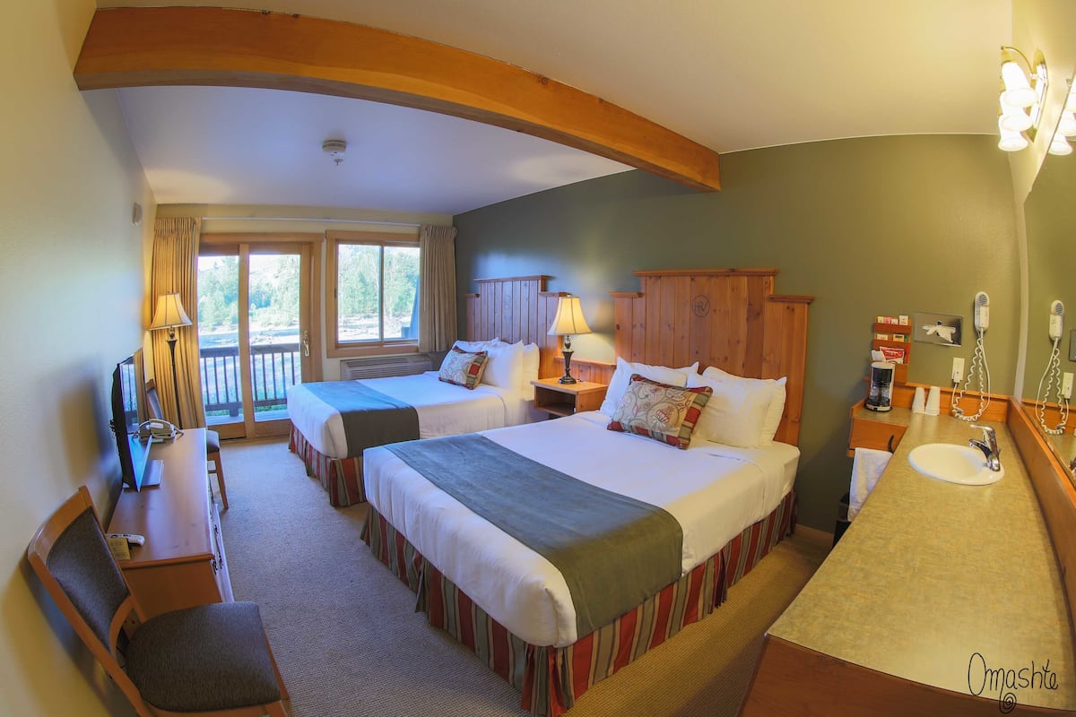 A hotel room with two beds, a desk with a chair, a desk lamp, a telephone, a sink with a mirror, and a view of the outdoors through a sliding door.