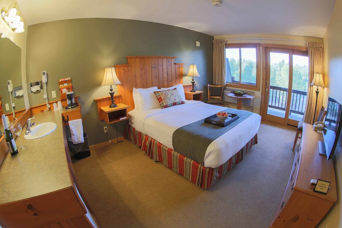 A hotel room with a large bed, a wooden headboard, a small table with a lamp, a window with curtains, and a bathroom with a sink and mirror.