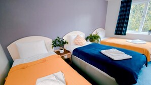 Habitación de hotel con dos camas, cada una con una colcha naranja y almohadas blancas. Se ve una ventana con vistas a árboles y una cortina azul.