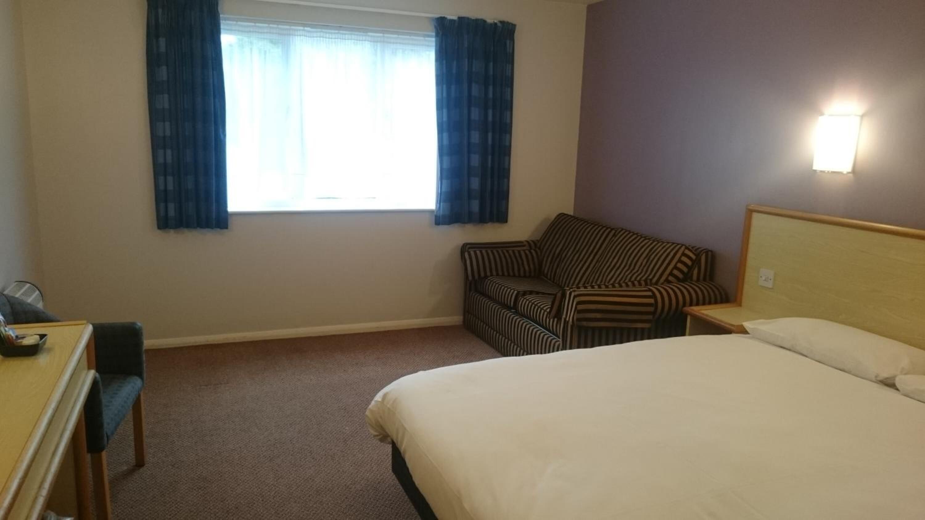 Une chambre d’hôtel avec un lit, un fauteuil à rayures, un bureau avec une chaise et une fenêtre avec des rideaux.