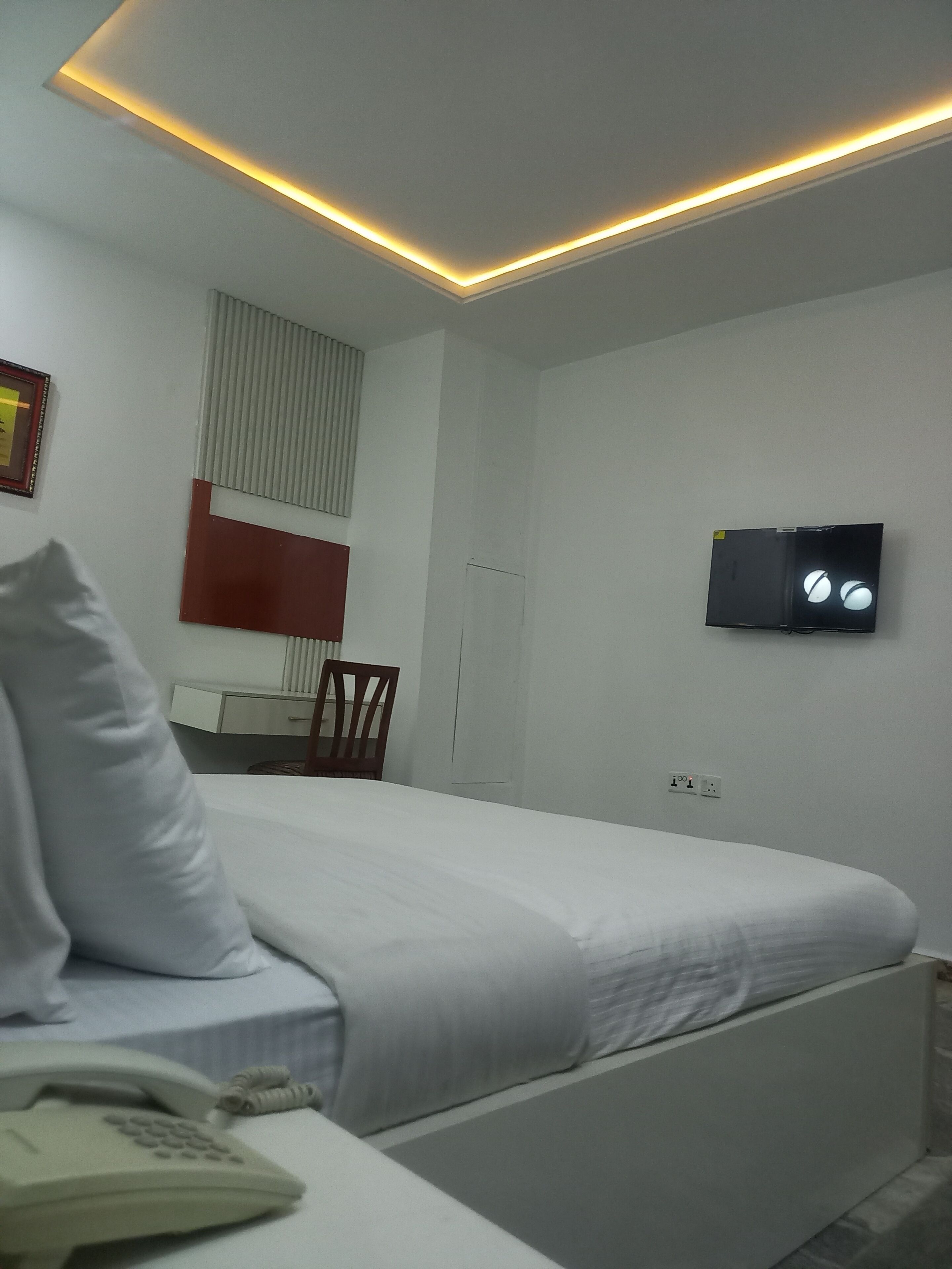 Une chambre d’hôtel comprenant un lit, un téléphone sur la table de chevet, un téléviseur fixé au mur, une fenêtre avec des stores et une petite table avec une chaise.