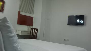 A hotel room with a bed, a bedside phone, a TV mounted on the wall, a window with blinds, and a small table with a chair.