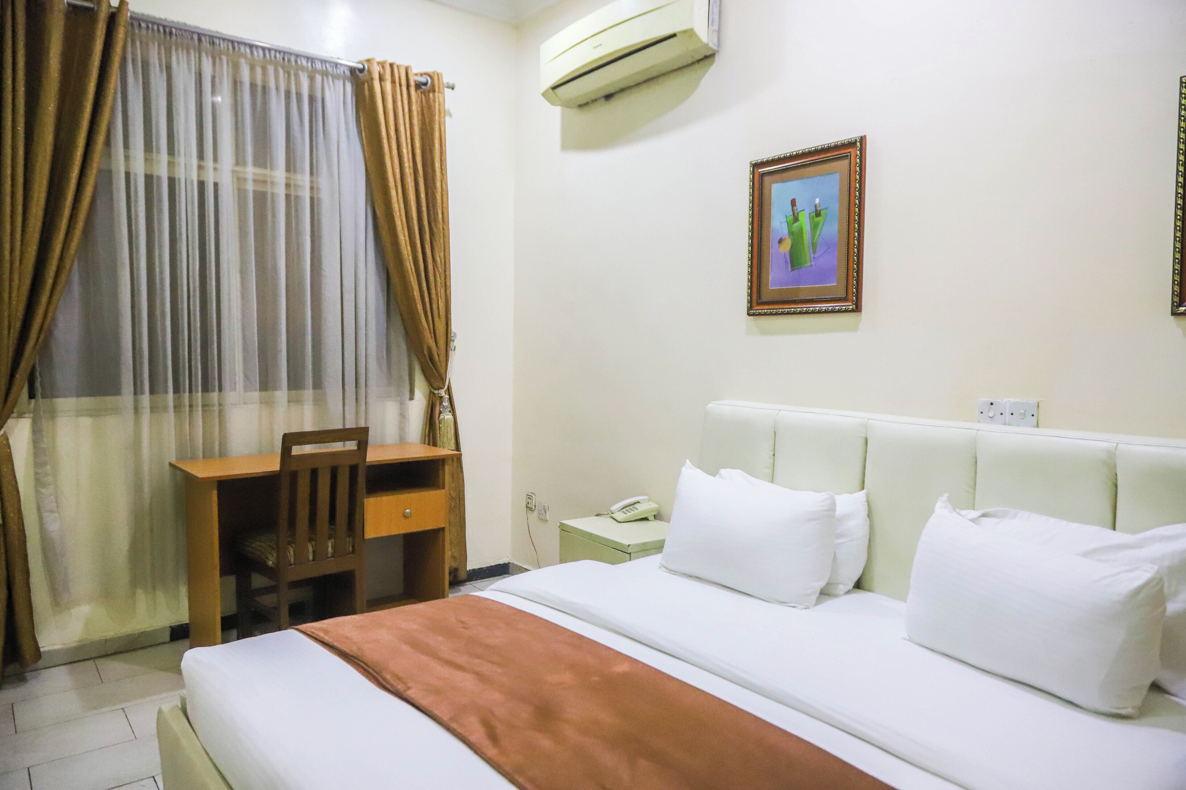 A hotel room with a bed, a desk, a chair, an air conditioner, and a framed picture on the wall.