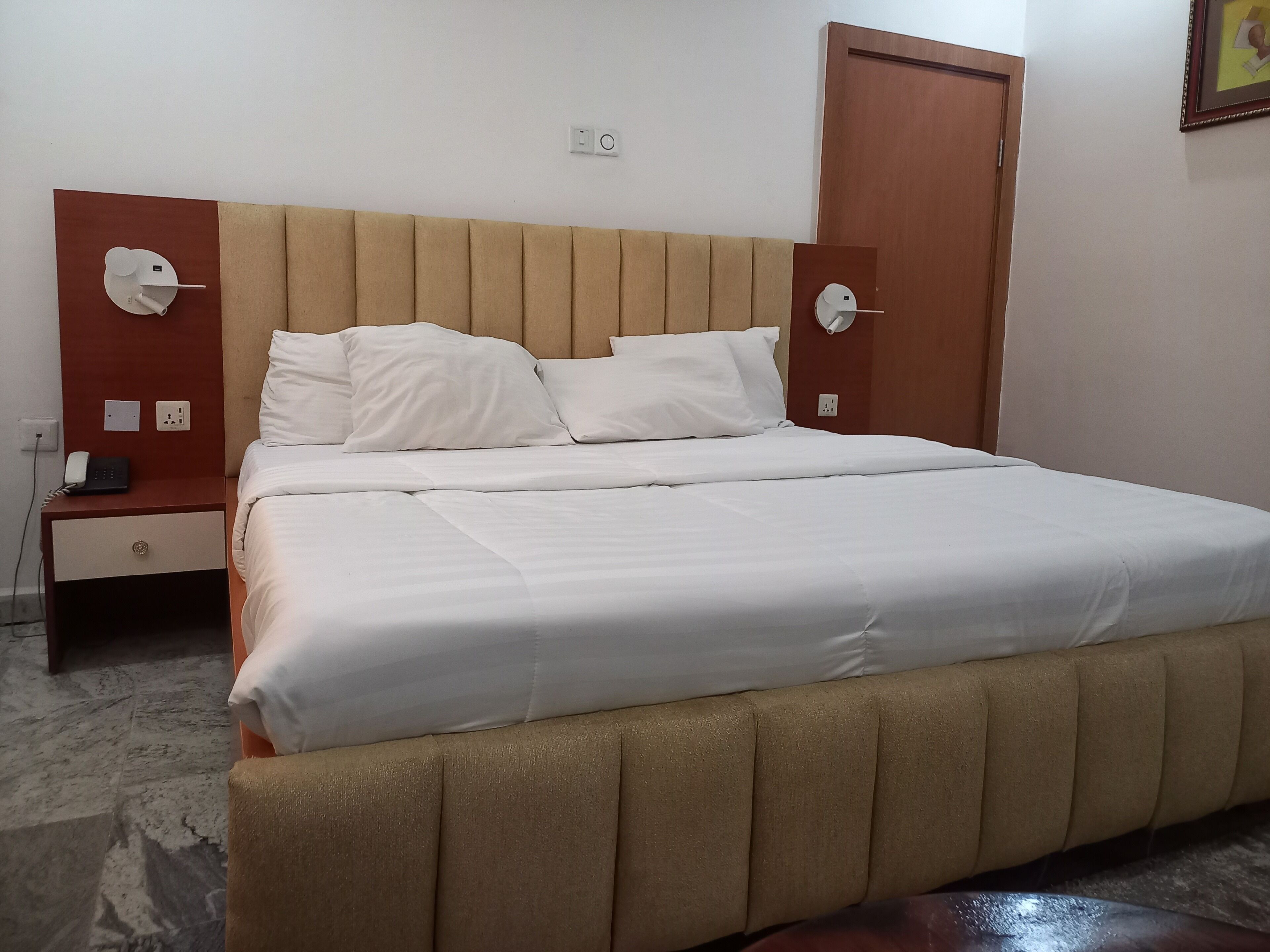 A neatly made bed with white linens, a wooden headboard, and a nightstand with a phone and a power outlet.