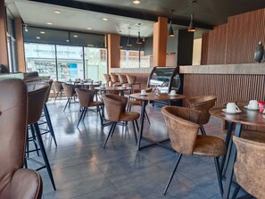 Interior moderno de cafetería con sillas y mesas de cuero, ventanas amplias y una máquina de café.