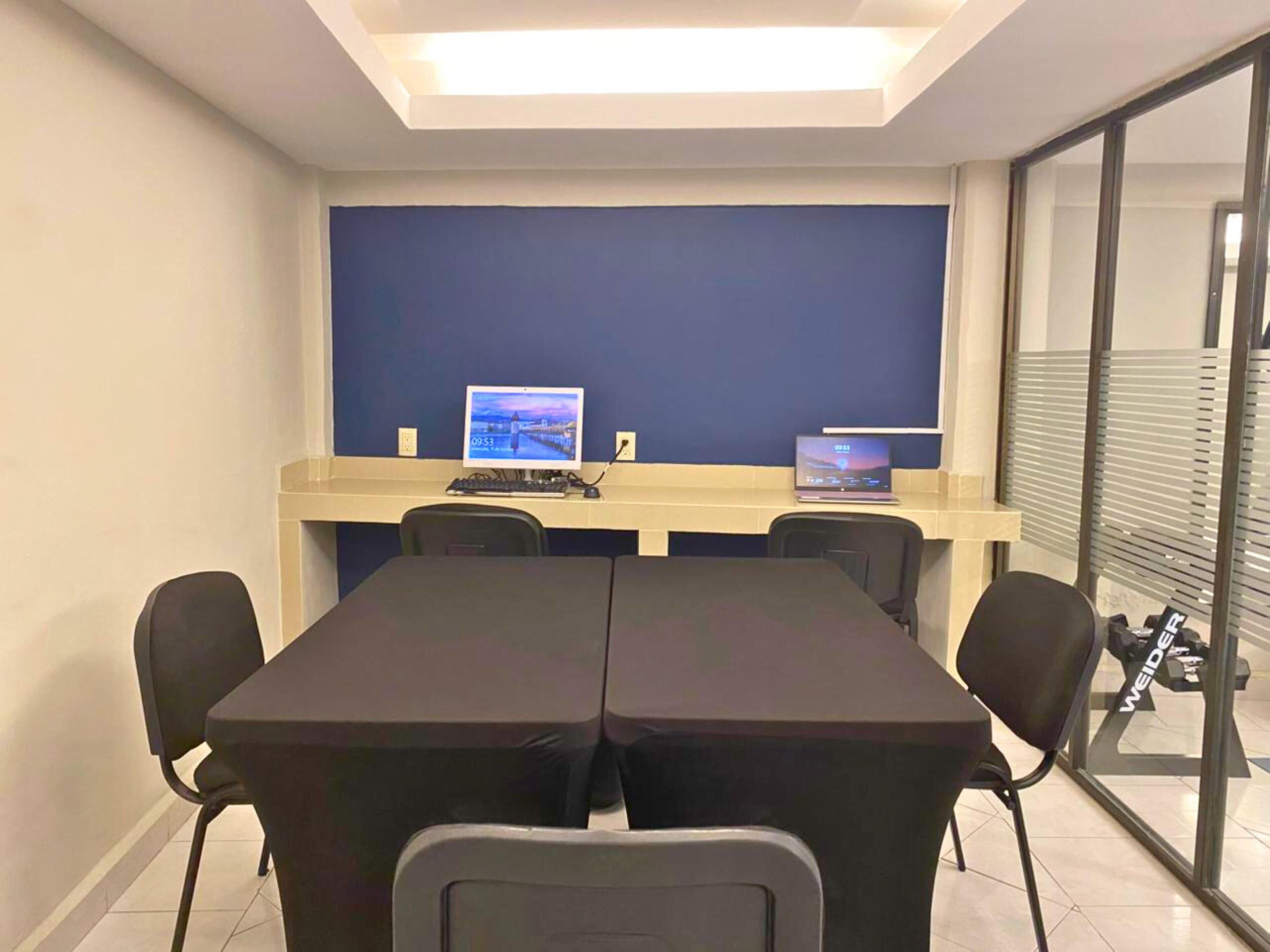 Un salón de conferencias con una mesa larga, dos computadoras y un ventanal con persianas.