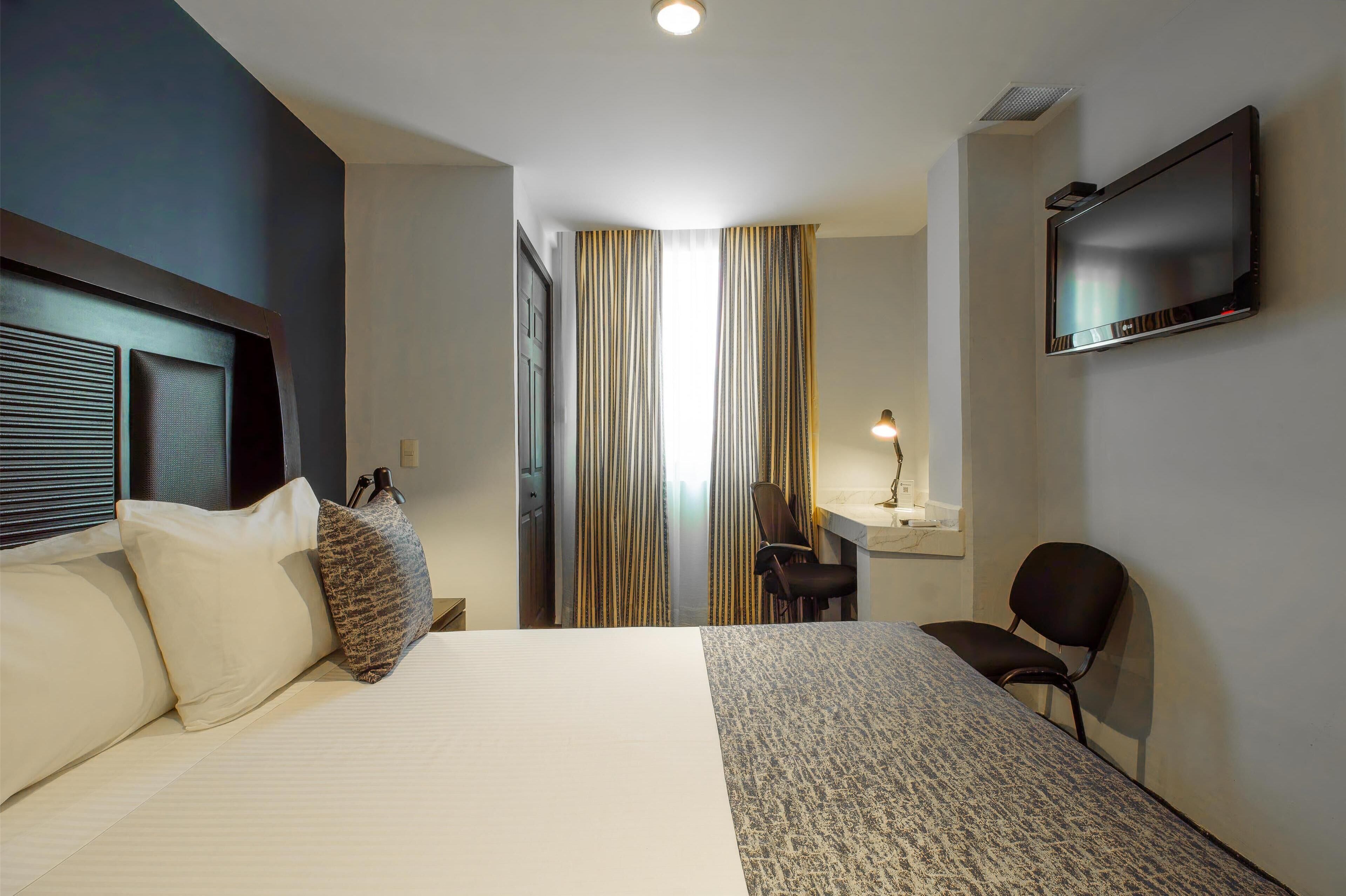 Quarto de hotel com uma cama grande, uma escrivaninha com cadeira, televisão na parede e janela com cortinas.