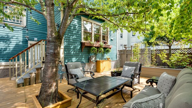 A wooden deck with outdoor furniture, a tree, and a house with a window and planter. - O Canada House (Vancouver)