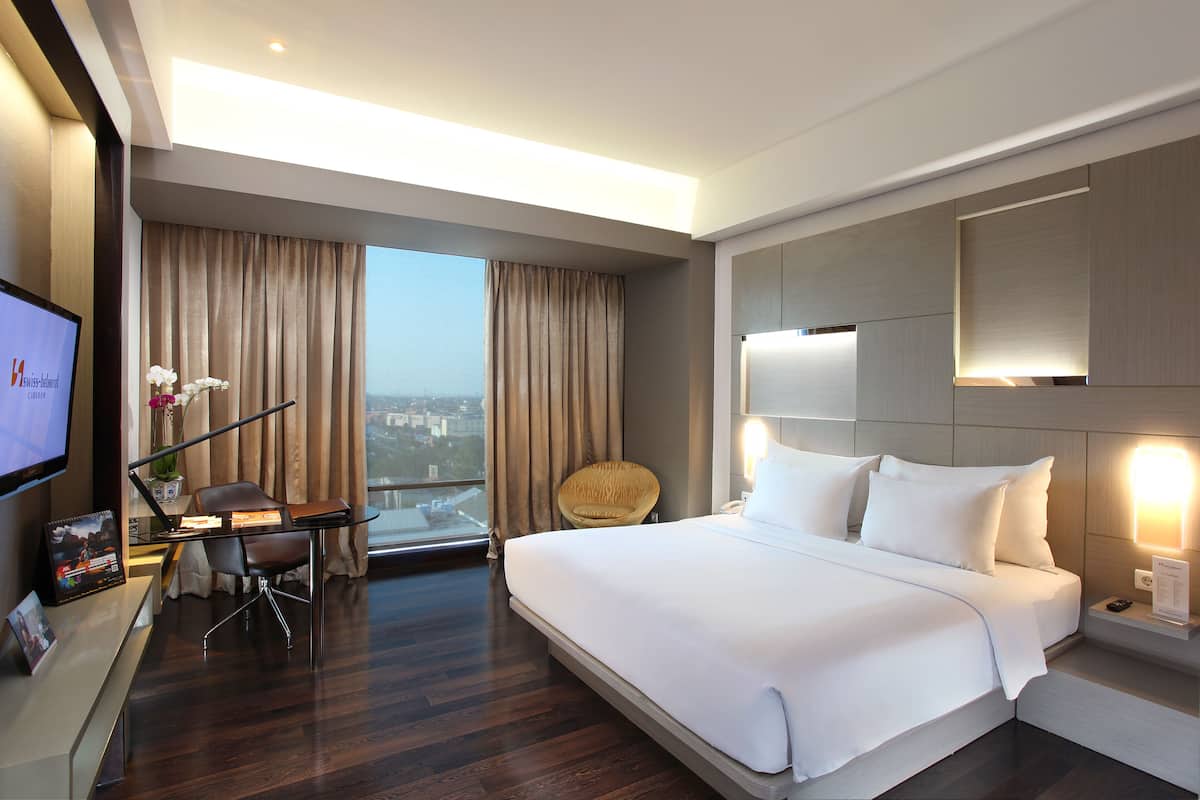 Habitación de hotel moderna con una cama grande, un escritorio con silla, televisión y vista a la ciudad.