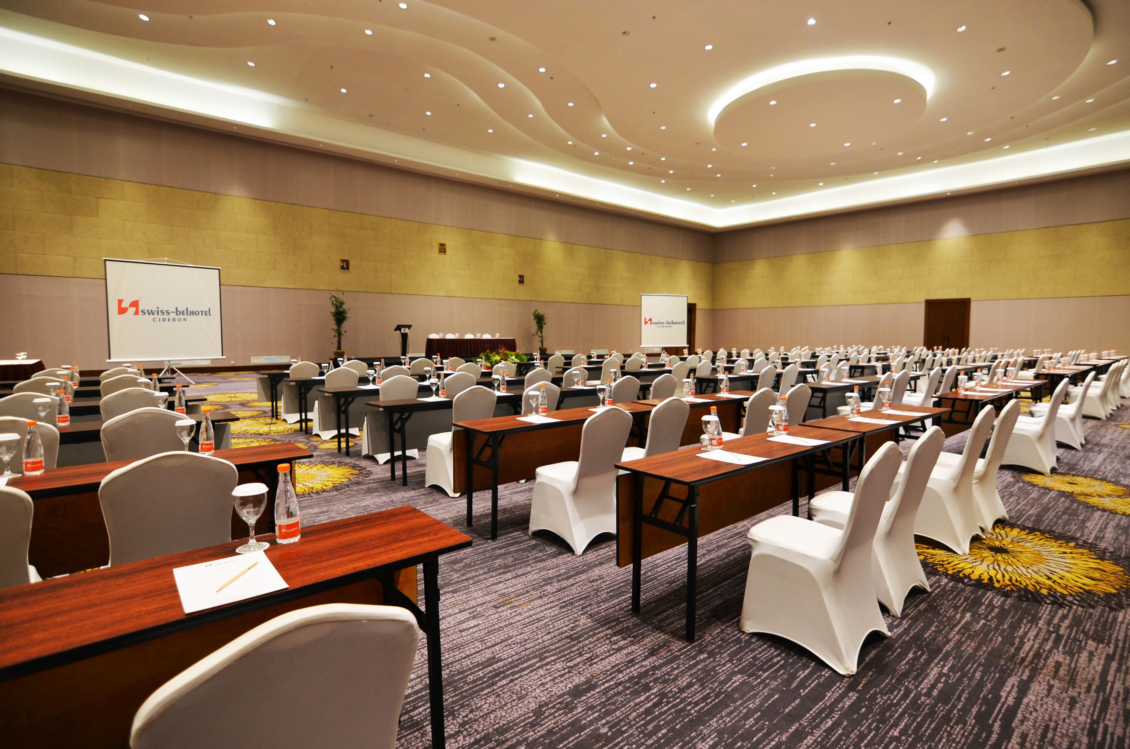 A conference room with rows of tables and chairs, a stage with a podium, and presentation screens.