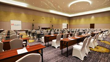 A conference room with rows of tables and chairs, a stage with a podium, and presentation screens.