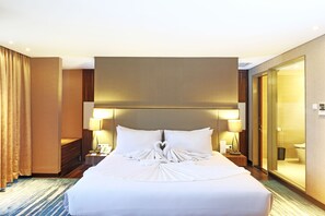 Habitación de hotel con una cama bien hecha, dos lámparas de noche, un espejo grande y un baño con paredes de vidrio.