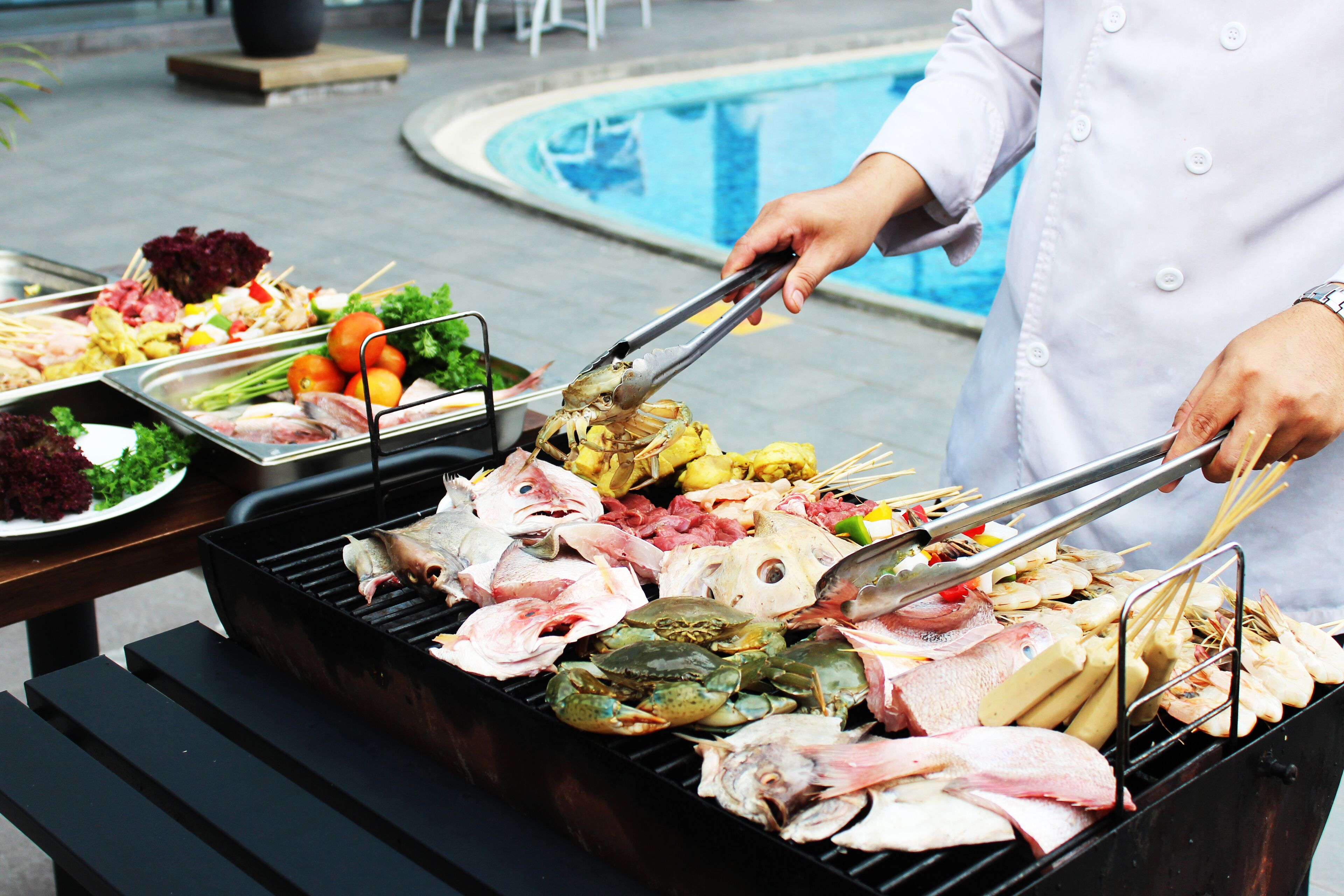 Een barbecue met diverse vleessoorten en zeevruchten, een zwembad op de achtergrond en iemand die aan het grillen is.