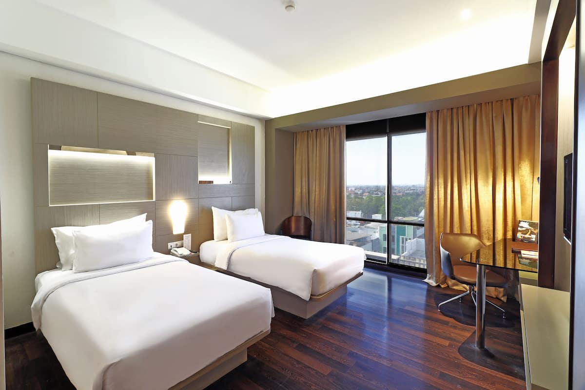 Habitación de hotel con dos camas, un escritorio, una silla y un amplio ventanal con vistas a la ciudad.
