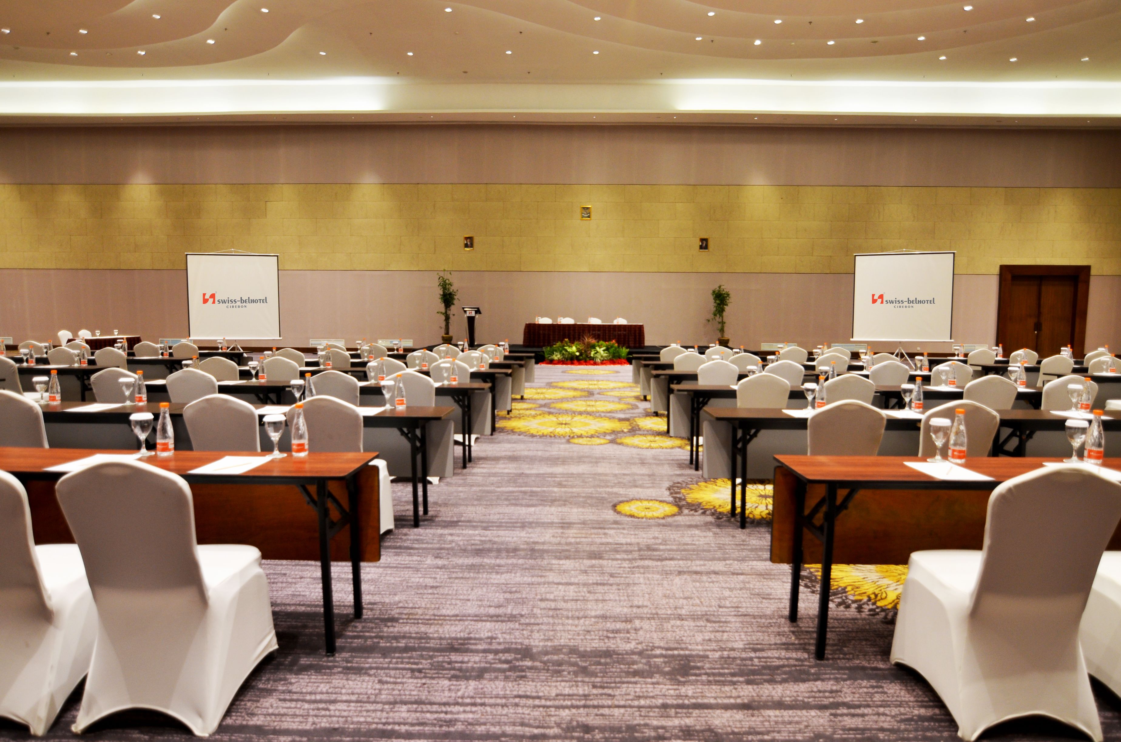 A conference room with rows of tables and chairs, a carpeted floor, and two projection screens with the logo "Swiss-Belhotel".