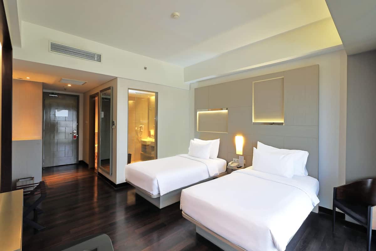 Habitación de hotel con dos camas, un escritorio, una silla, un baño con espejo y una puerta que da a otra habitación.