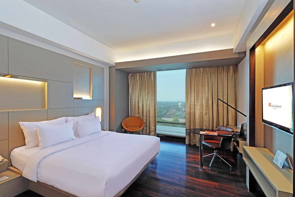 A hotel room with a bed, desk, chair, and a large window with curtains.